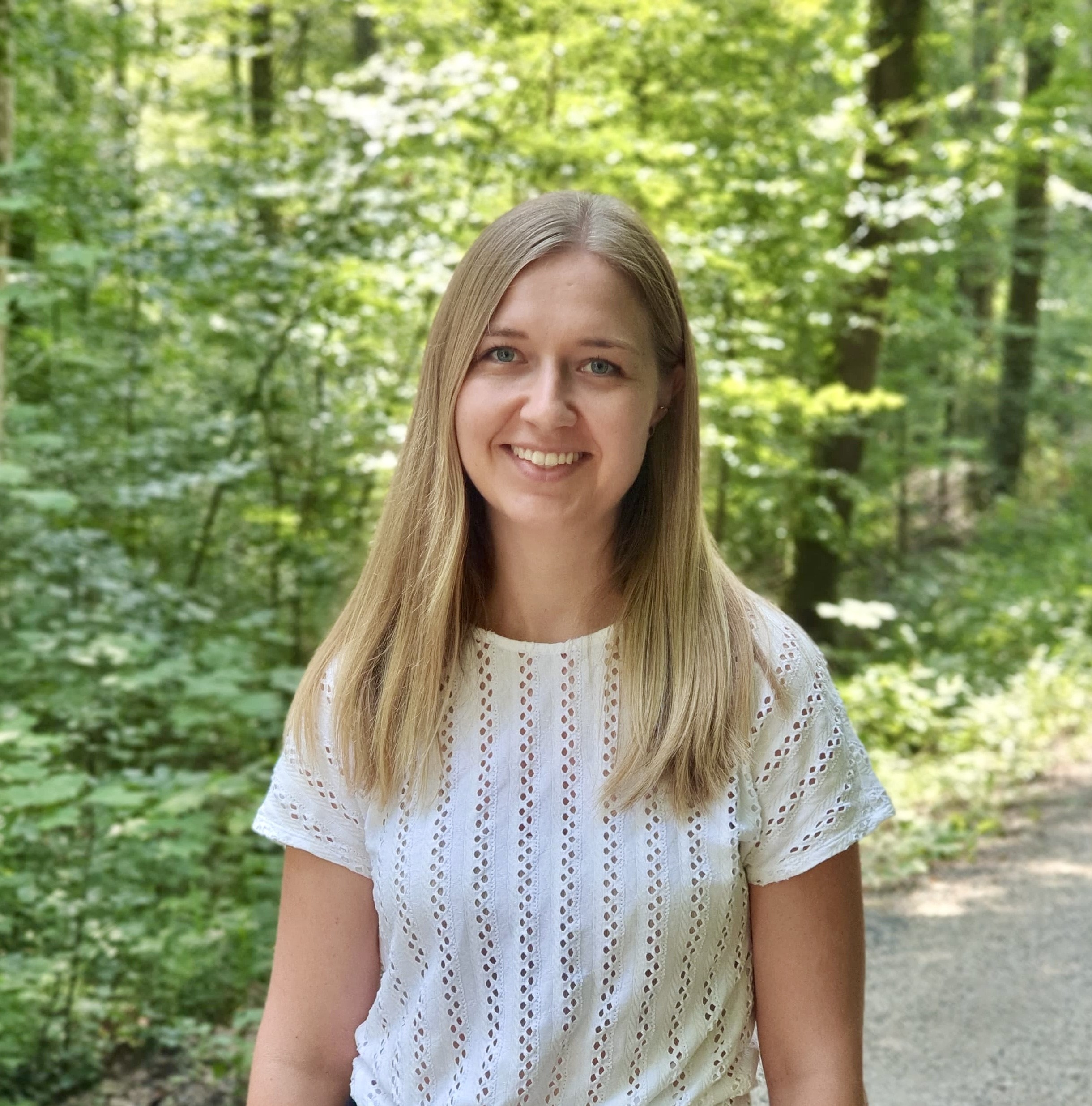 image01 Eine Frau mit blonden langen Haaren und einem weißen T-Shirt. Im Hintergrund sind grüne Bäume.